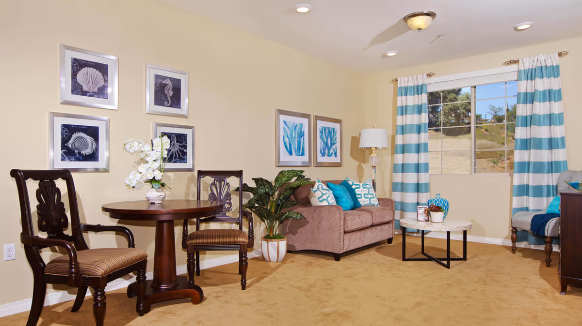 A cozy living room with beige walls and carpet, featuring a brown loveseat with blue and white patterned pillows, a round marble coffee table with decorative items, a floor lamp, and a window with blue and white striped curtains. There is also a small round wooden table with two wooden chairs, a potted plant, and framed ocean-themed artwork on the walls.