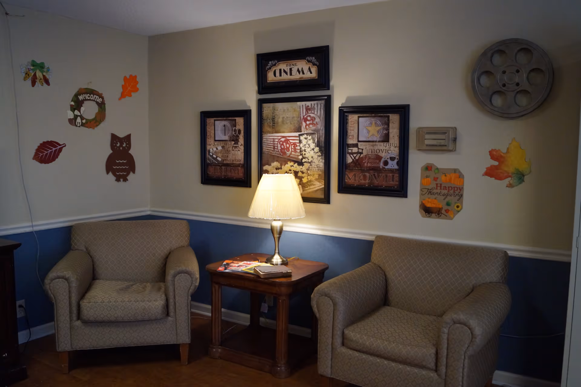 A cozy corner in a senior living facility with two beige patterned armchairs separated by a wooden side table holding a lit lamp and some magazines. The wall behind features framed movie-themed artwork, a large decorative film reel, and autumn-themed decorations including leaves, an owl, and a 'Happy Thanksgiving' sign.