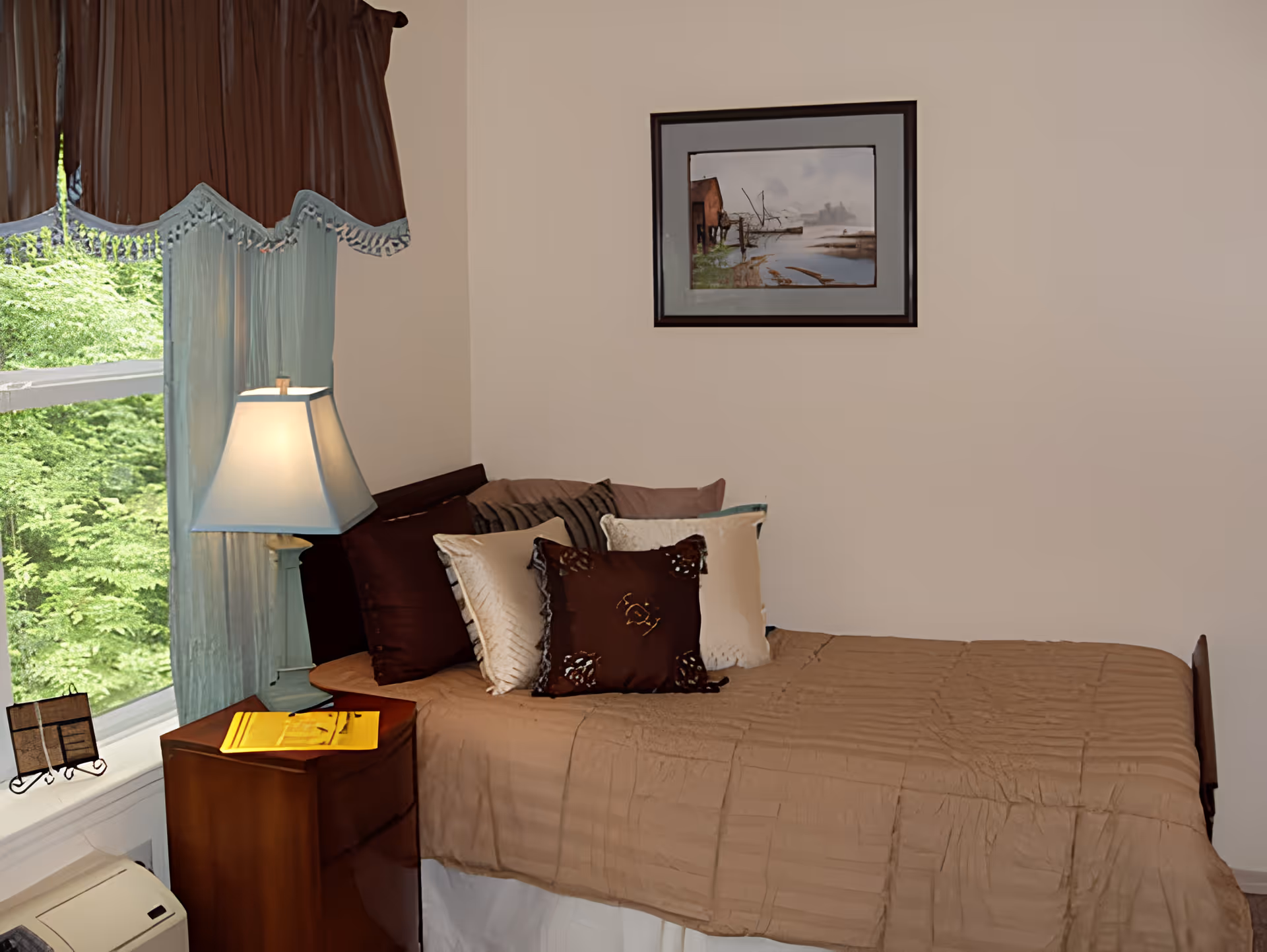 A small bedroom with a single bed covered in a brown quilt and multiple decorative pillows. Next to the bed is a wooden nightstand with a lamp and a yellow book or folder on it. A window with blue curtains and a brown valance lets in natural light, and a framed painting of a waterfront scene hangs on the beige wall above the bed.
