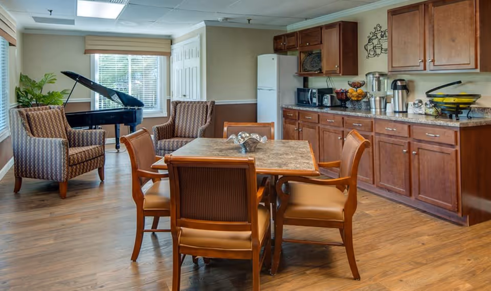 A cozy common area with a square table surrounded by four wooden chairs with cushioned seats. Behind the table is a kitchenette with wooden cabinets, a refrigerator, microwave, coffee maker, and various kitchen items on the countertop. To the left, there are two patterned armchairs and a black grand piano near a window with blinds and a beige valance. The room has wood flooring and neutral-colored walls.
