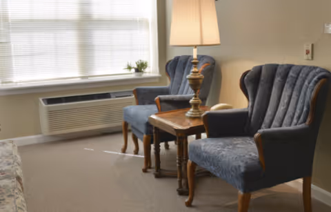 A cozy sitting area with two blue upholstered armchairs and a wooden side table between them. A brass table lamp with a beige shade is on the side table. There is a window with closed blinds and a small potted plant on the windowsill. The room has beige walls and carpeted floor.