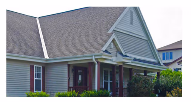 Exterior view of a residential-style building with gray siding, a steeply pitched roof, and a covered porch supported by columns. There are shrubs and greenery in front of the building.