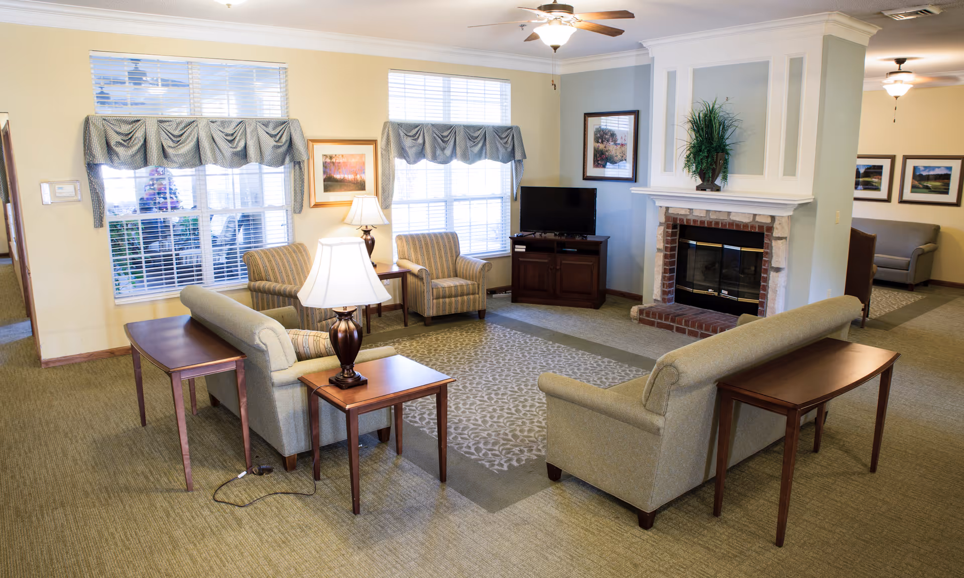 A cozy living room area in a senior living facility featuring two sofas facing each other with wooden side tables and lamps, two striped armchairs near large windows with valances, a patterned area rug, a brick fireplace with a plant on the mantel, a flat-screen TV on a wooden cabinet, and framed artwork on the walls.