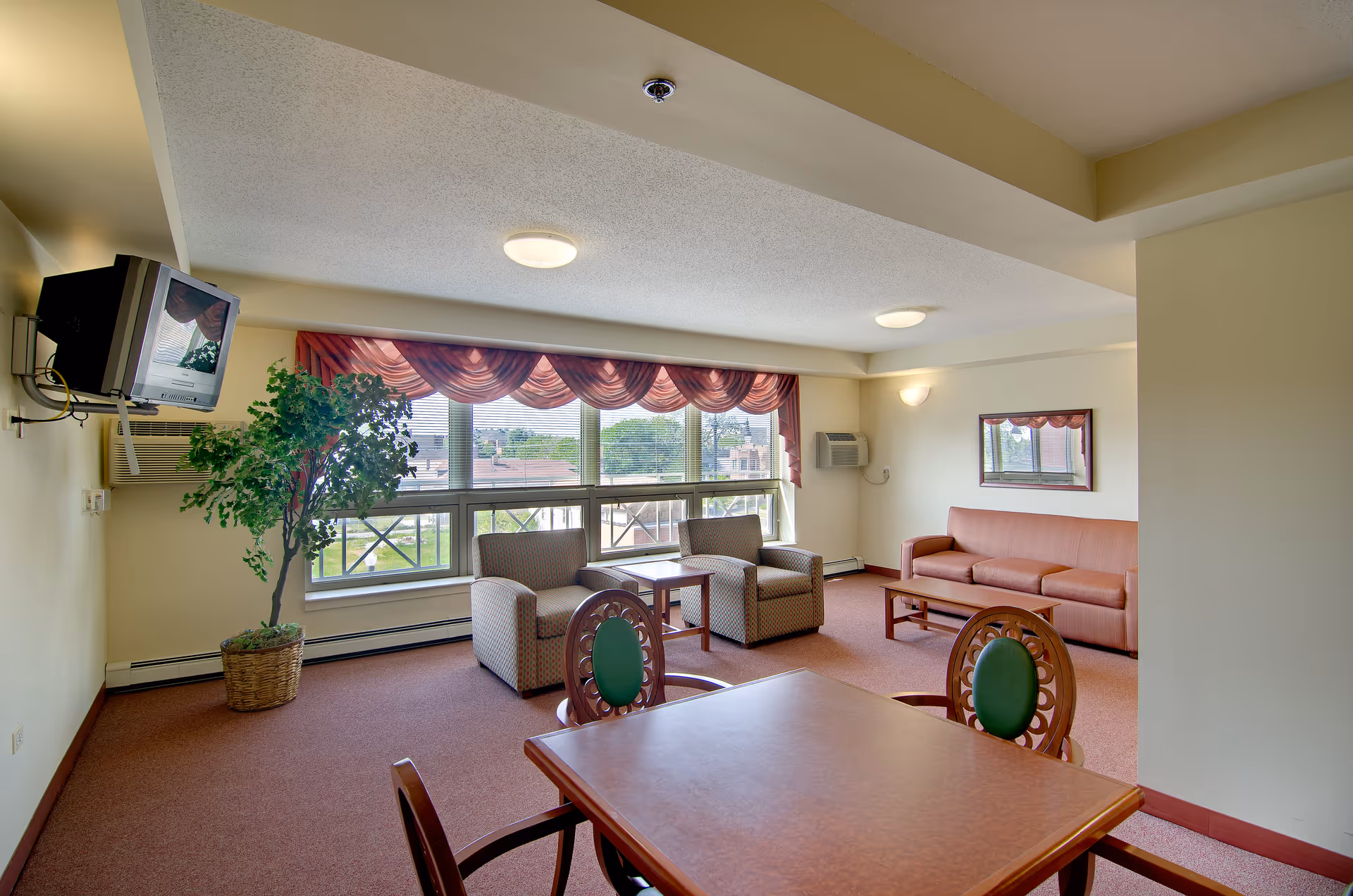 Well-lit senior living common room with seating, a table, TV, and large windows.