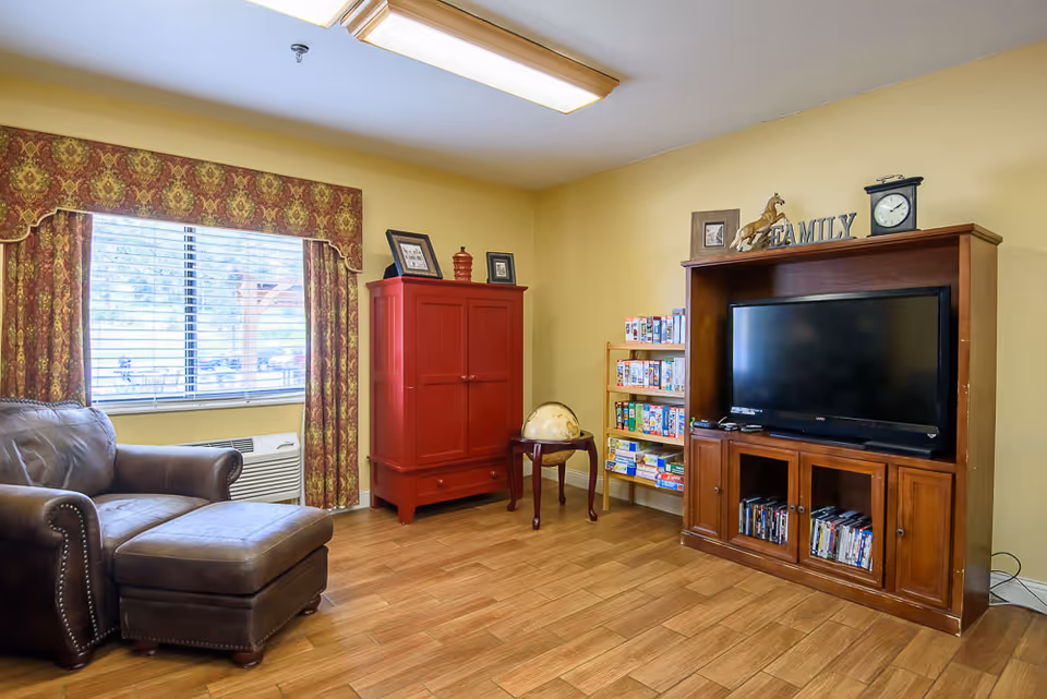 A cozy living room with a brown leather armchair and matching ottoman near a window with patterned curtains. A red cabinet stands against the wall next to a small table holding a globe. A wooden entertainment center houses a flat-screen TV, DVDs, and decorative items including a clock, a horse figurine, and a 'FAMILY' sign. A small shelf with board games is also visible.
