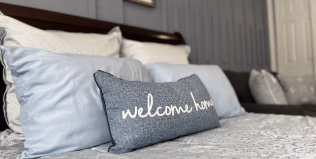 Close-up view of a bed with multiple pillows, including a decorative pillow with the words 'welcome home' embroidered on it, in a softly lit bedroom.