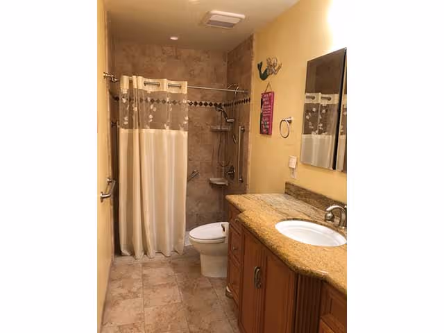 A bathroom with beige tiled floor and walls, featuring a shower area with a curtain, a white toilet, and a wooden vanity with a granite countertop and a sink. A mirror is mounted above the sink, and there are grab bars near the shower and toilet for accessibility.