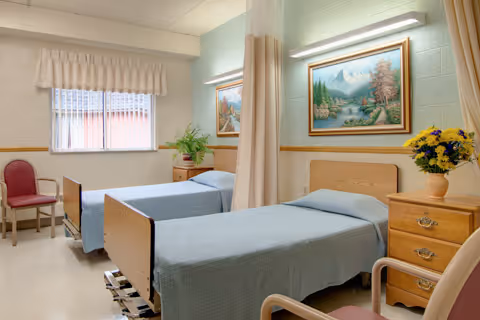 A bright and clean room with two single beds covered with light blue bedspreads, separated by a beige privacy curtain. There is a wooden nightstand with a vase of yellow flowers, a framed landscape painting on the wall, a window with white blinds and valance, and two chairs placed near the beds.