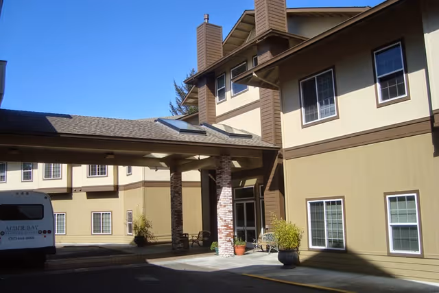 Exterior view of Alder Bay Assisted Living facility showing a covered entrance supported by brick columns, beige and brown building walls with multiple windows, potted plants near the entrance, and a parked van with Alder Bay logo on the left side.