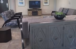A cozy common living area with a sofa and armchairs facing a TV on a console, a decorative cabinet behind the sofa, and framed artwork on the walls.