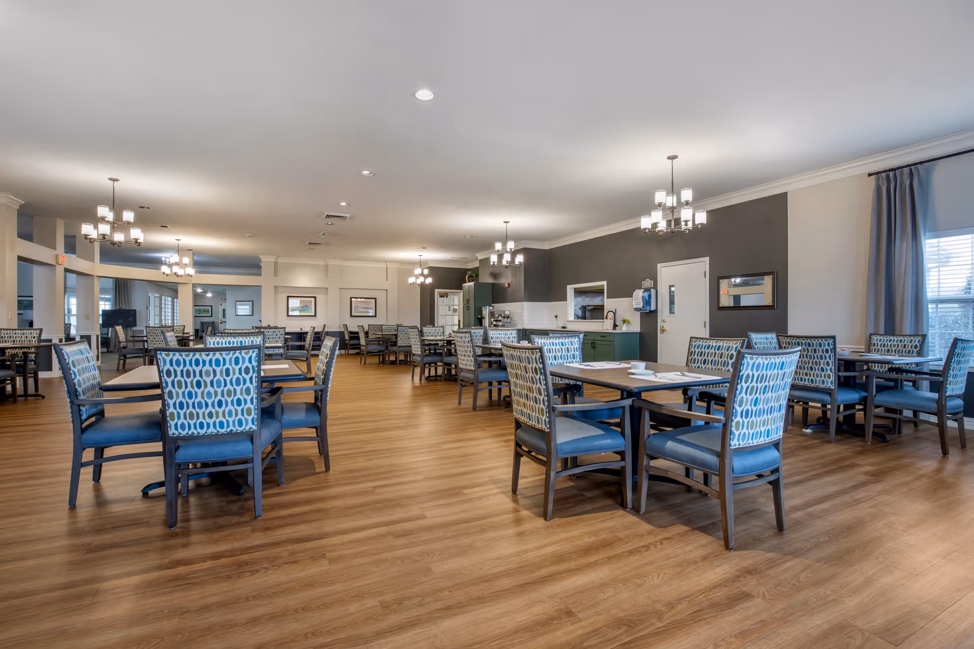 A spacious dining room with multiple tables and chairs arranged neatly. The chairs have blue patterned upholstery, and the room features wooden flooring, chandeliers hanging from the ceiling, and large windows with curtains allowing natural light to enter.