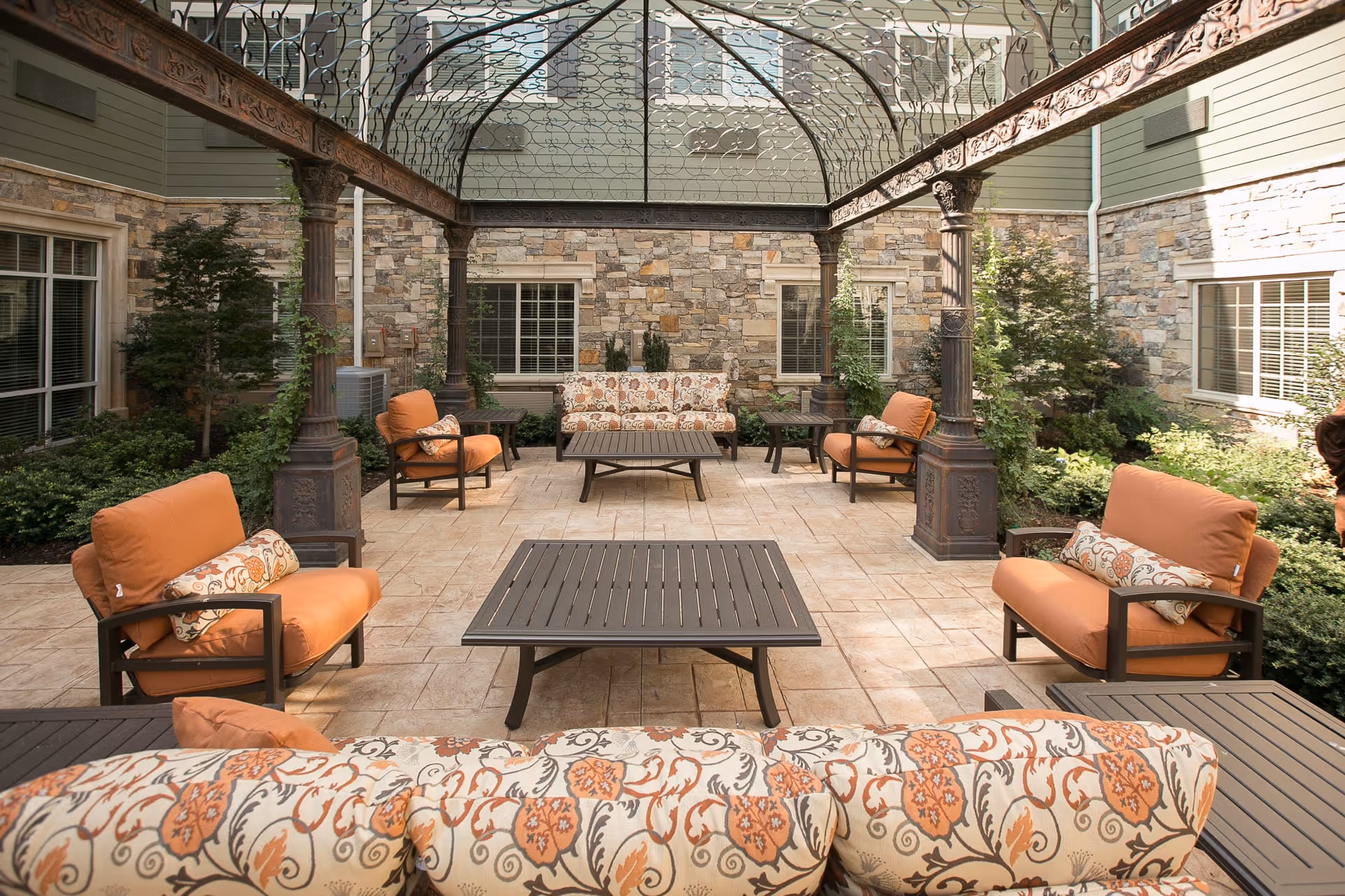 Outdoor patio area with cushioned seating including armchairs and sofas arranged around two rectangular coffee tables. The space is covered by a decorative metal pergola and surrounded by stone walls with windows and greenery.
