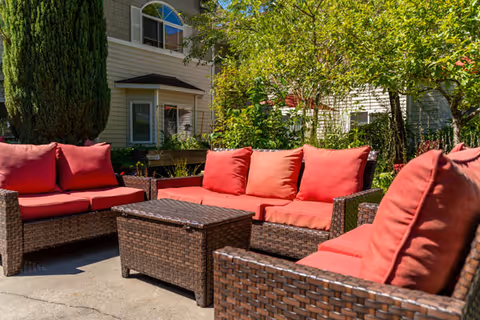 Wicker outdoor seating with red cushions arranged around a low table on a sunny patio courtyard in front of a residential building.