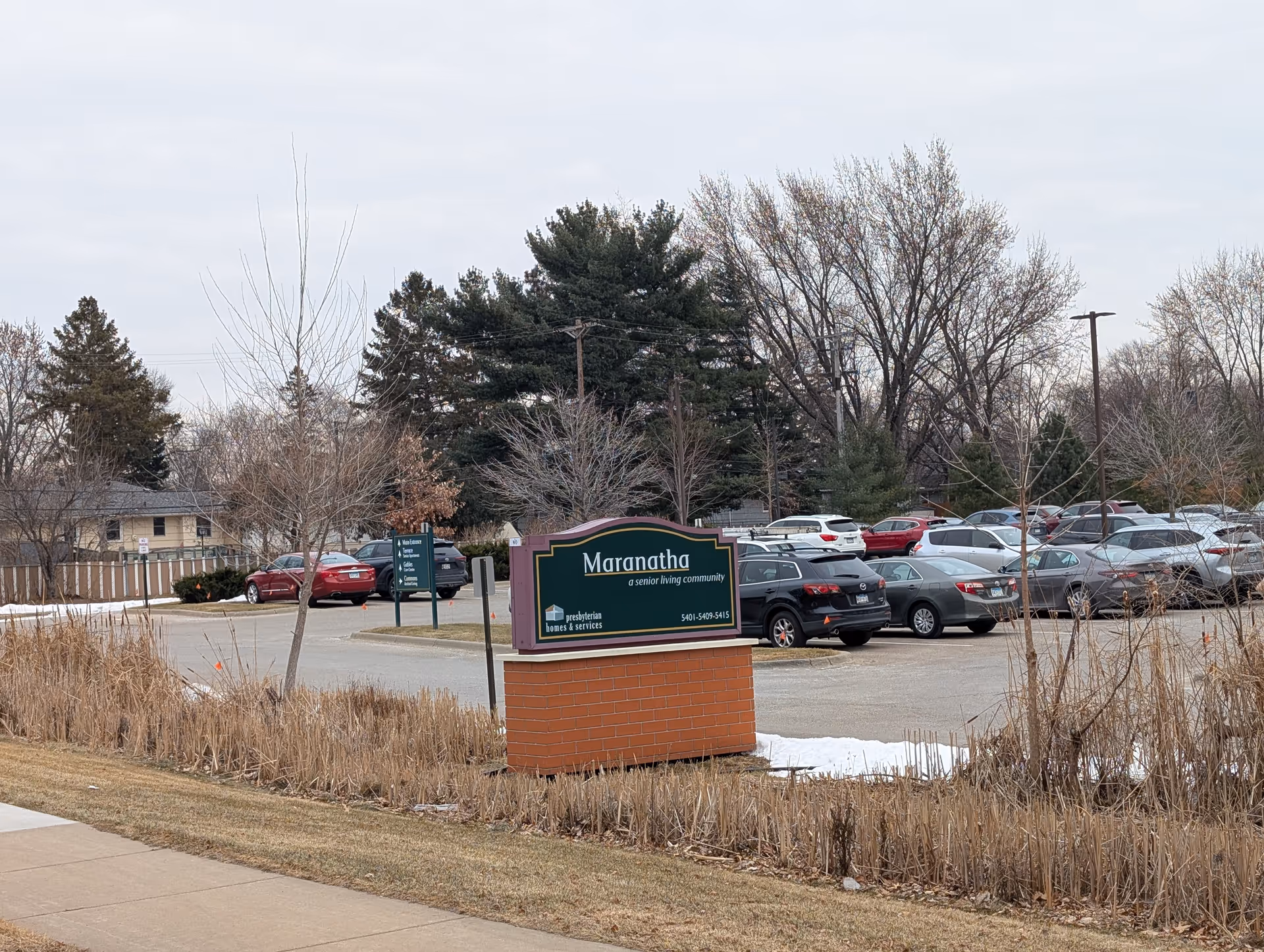 Outdoor view of the parking lot at Maranatha, a senior living community, with a sign in the foreground and several parked cars surrounded by trees and dry grass.