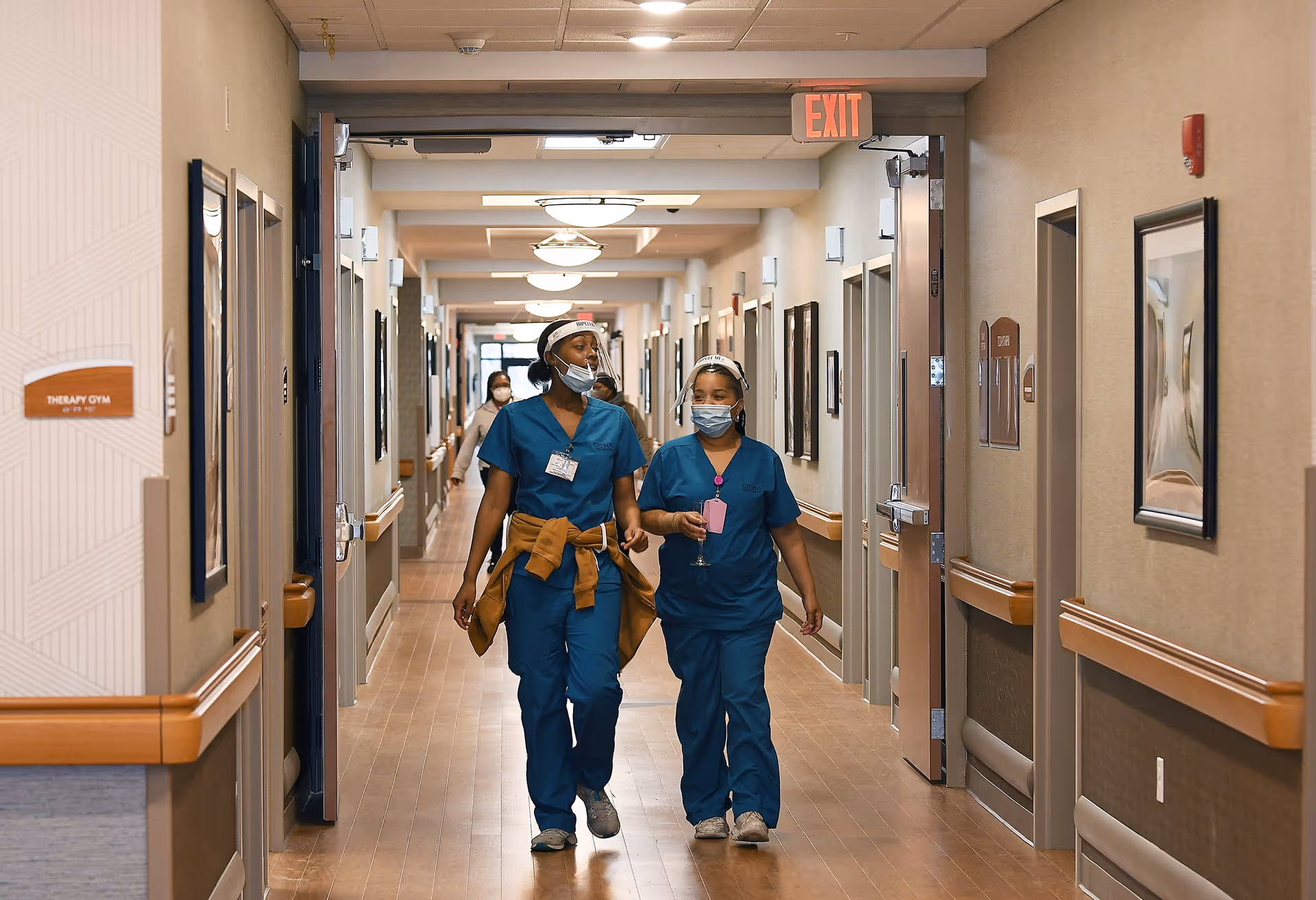 Two healthcare workers wearing blue scrubs and face masks walking down a well-lit hallway in a nursing and rehabilitation facility. The hallway has handrails on both sides, framed pictures on the walls, and several doors leading to different rooms. A sign on the left indicates a therapy gym, and an exit sign is visible at the end of the hallway.
