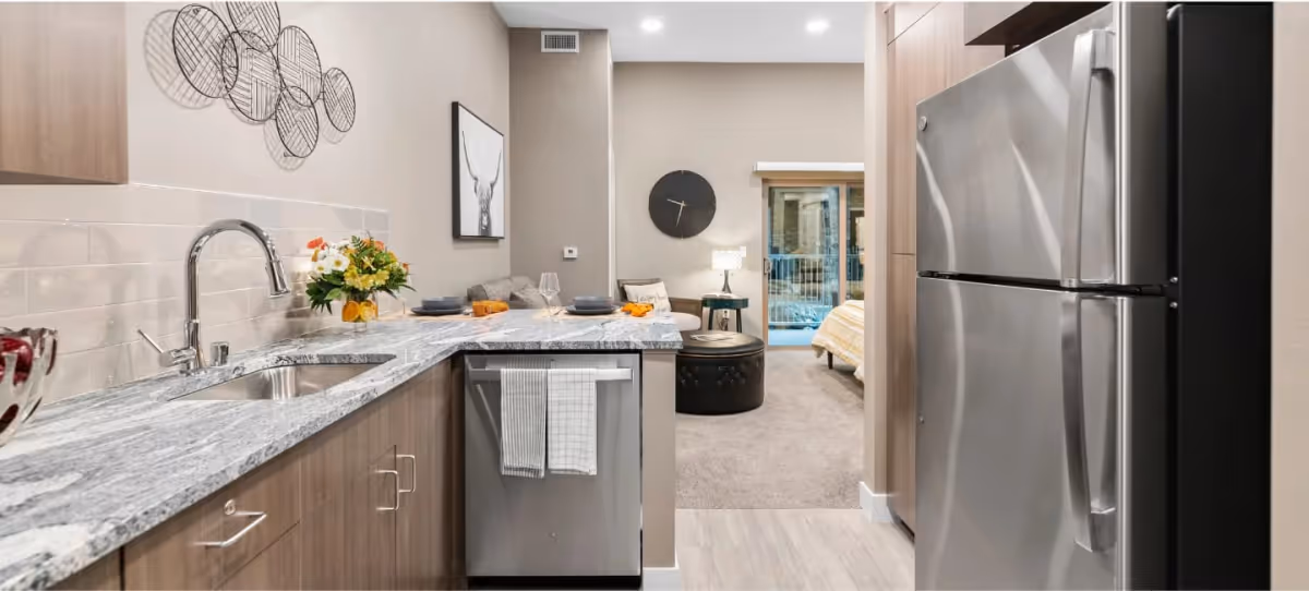Modern kitchen area with a marble countertop, stainless steel sink, dishwasher, and refrigerator. The kitchen opens into a living space with a sofa, round ottoman, wall clock, and a bedroom visible in the background with a bed and sliding glass door.