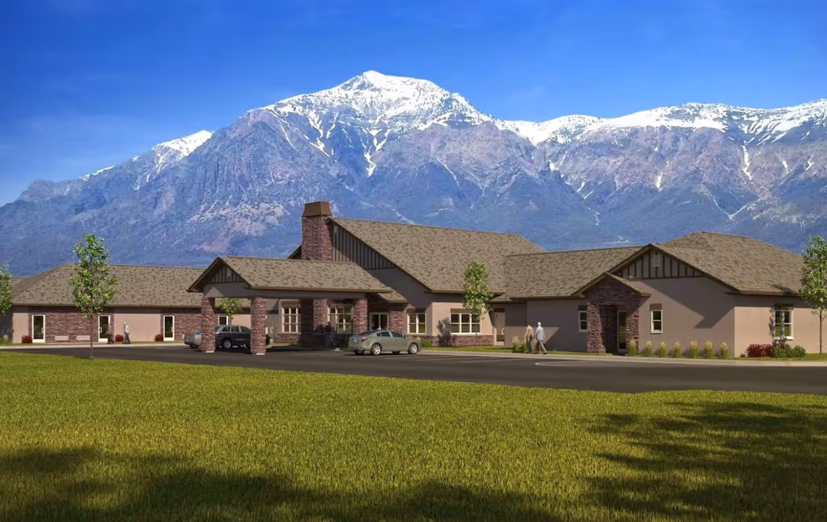 Exterior view of Quail Meadow Assisted Living facility with a large building featuring a covered entrance, parked cars, and a backdrop of tall snow-capped mountains under a clear blue sky.