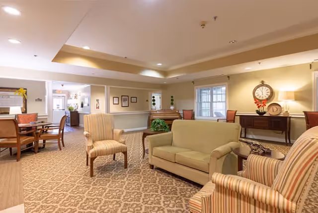 A cozy senior living facility common area with patterned carpet, beige walls, and recessed lighting. The room features a green loveseat, two striped armchairs, a wooden side table with a lamp, a console table with a clock and decorative items, and a piano in the background. There is also a dining area with wooden chairs and tables visible to the left.