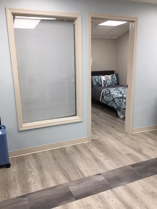 View of a hallway with light wood flooring and light blue walls, featuring a large window with frosted glass on the left and an open doorway on the right leading to a bedroom with a bed dressed in patterned blue and gray bedding.