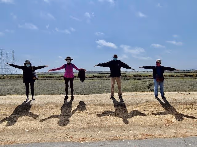 Four people standing outdoors on a dirt path with arms outstretched, wearing hats and face masks, casting long shadows on the ground under a partly cloudy sky.