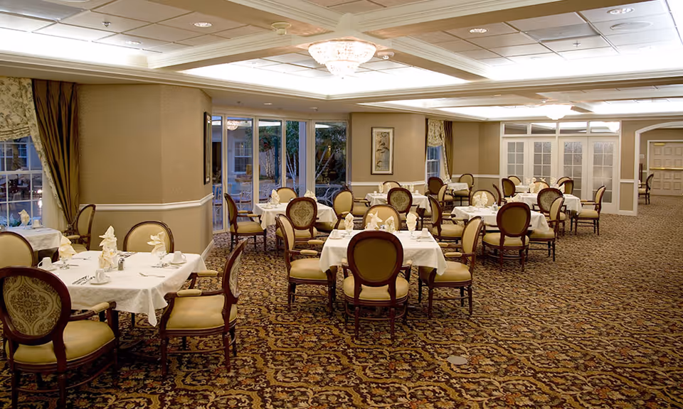 A spacious dining room with multiple round tables covered with white tablecloths, each set with napkins, cups, and silverware. The room features patterned carpet, upholstered chairs with wooden frames, large windows with curtains, and ceiling lights including chandeliers.
