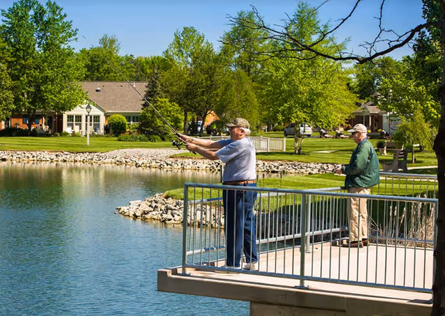 Two men fishing from a lakeside dock in a landscaped senior living community with houses and trees in the background.