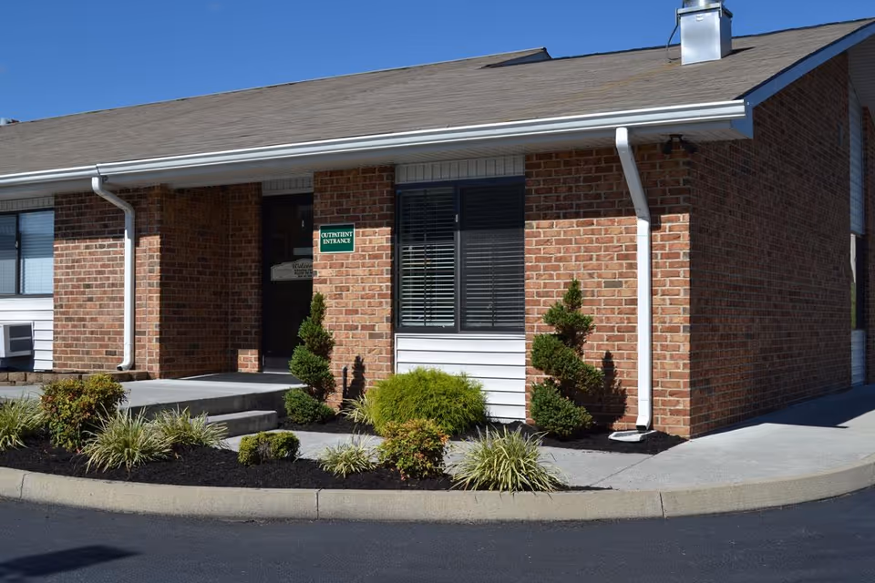 Brick single-story building front showing a covered entrance, windows, small shrubs, and a sign for the outpatient entrance.