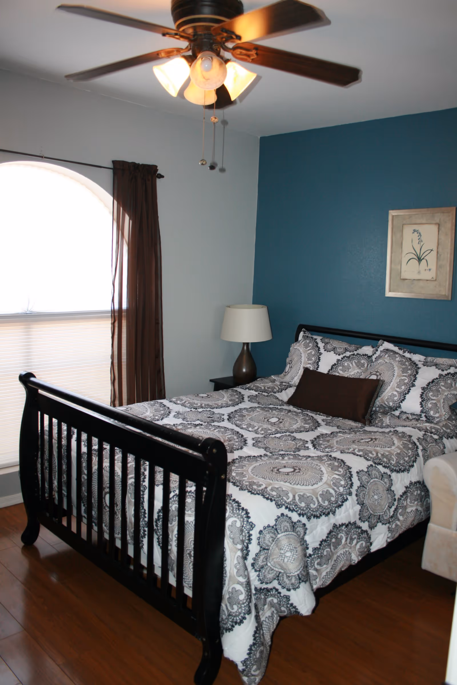A bedroom with a black wooden bed frame and a bed made with white and gray patterned bedding. There is a brown rectangular pillow on the bed. The room has wooden flooring, a blue accent wall with a framed botanical picture, a window with brown curtains, a ceiling fan with lights, and a bedside table with a lamp.
