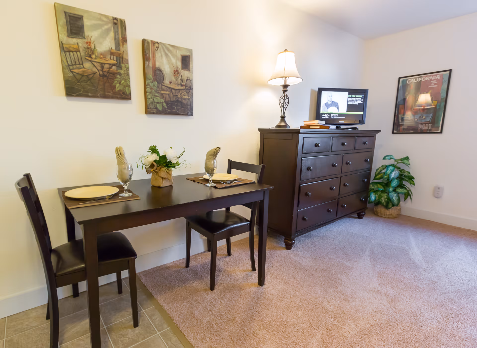 A small dining area with a dark wooden table set for two, including plates, glasses, and napkins. Two matching chairs are placed at the table. On the wall above the table are two paintings depicting outdoor café scenes. To the right, there is a dark wooden dresser with multiple drawers, a table lamp, a small TV, and some books on top. A framed California poster hangs on the wall above a green potted plant in a woven basket. The floor is carpeted except for the area under the table, which has tile flooring.