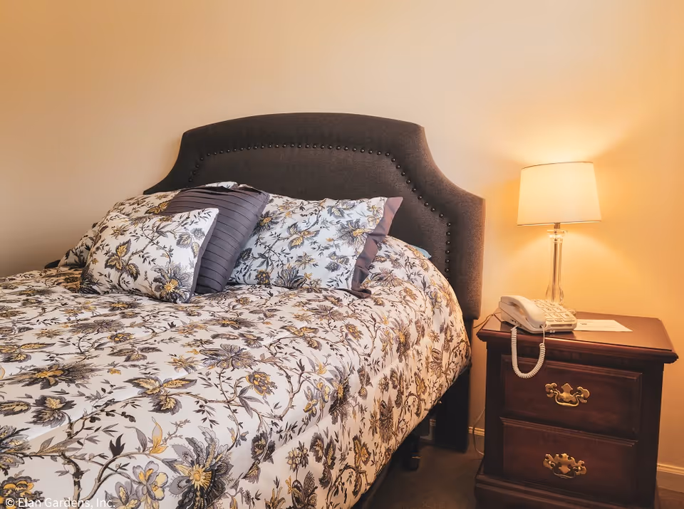 A neatly made bed with floral bedding and a gray upholstered headboard next to a wooden nightstand with a lamp and landline phone.