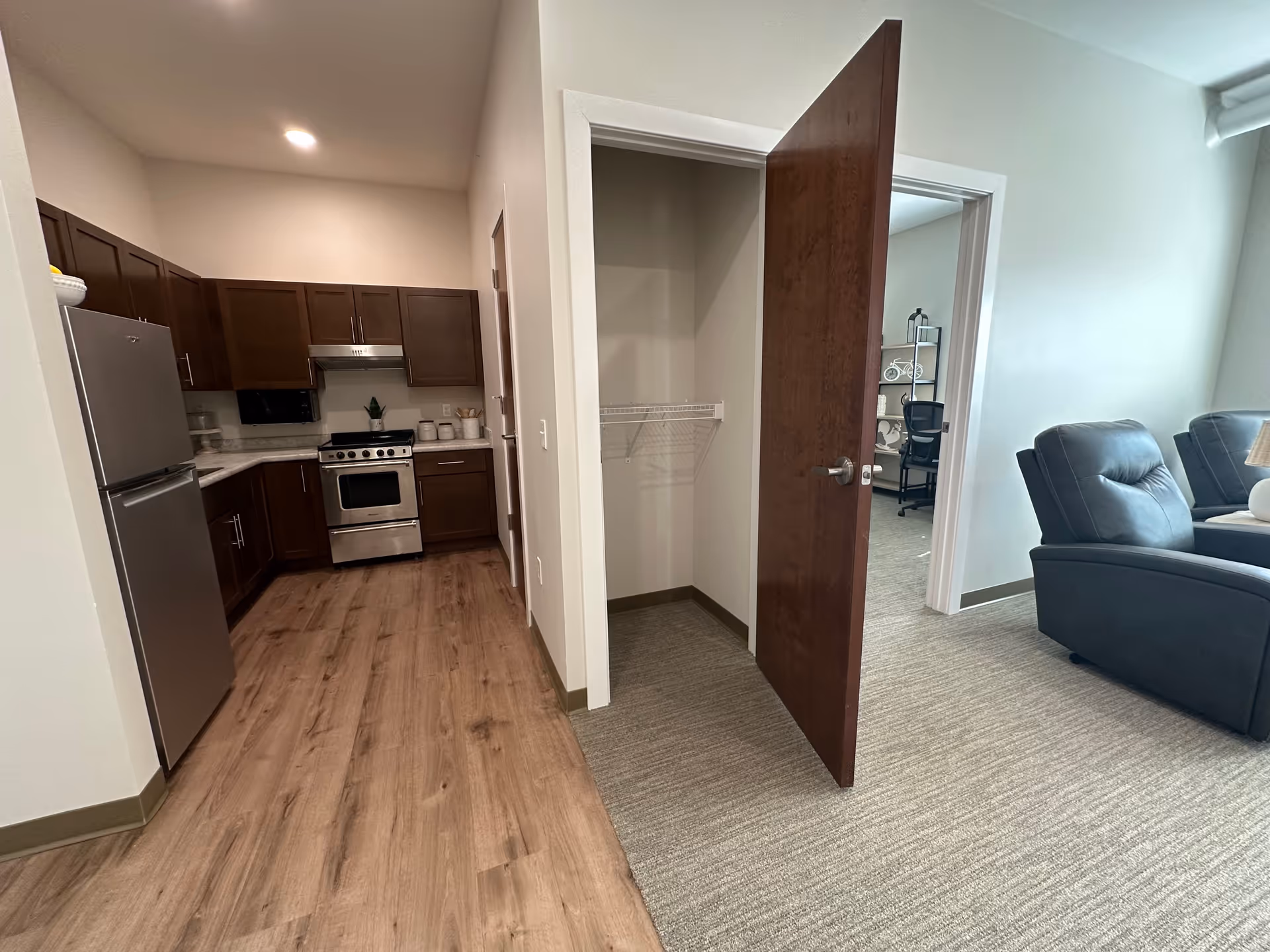 Interior view of a senior living community apartment showing a kitchen with wooden cabinets, stainless steel refrigerator and stove, wood flooring, an open closet with a wooden door, and part of a living area with carpeted floor and two dark-colored recliners.