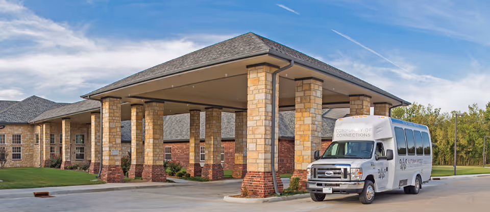 Exterior view of a senior living facility with a large covered entrance supported by stone pillars. A white shuttle bus labeled 'Community of Connections' is parked under the covered area. The building has a combination of stone and brick exterior with multiple windows and a well-maintained lawn. Trees and a blue sky with some clouds are visible in the background.