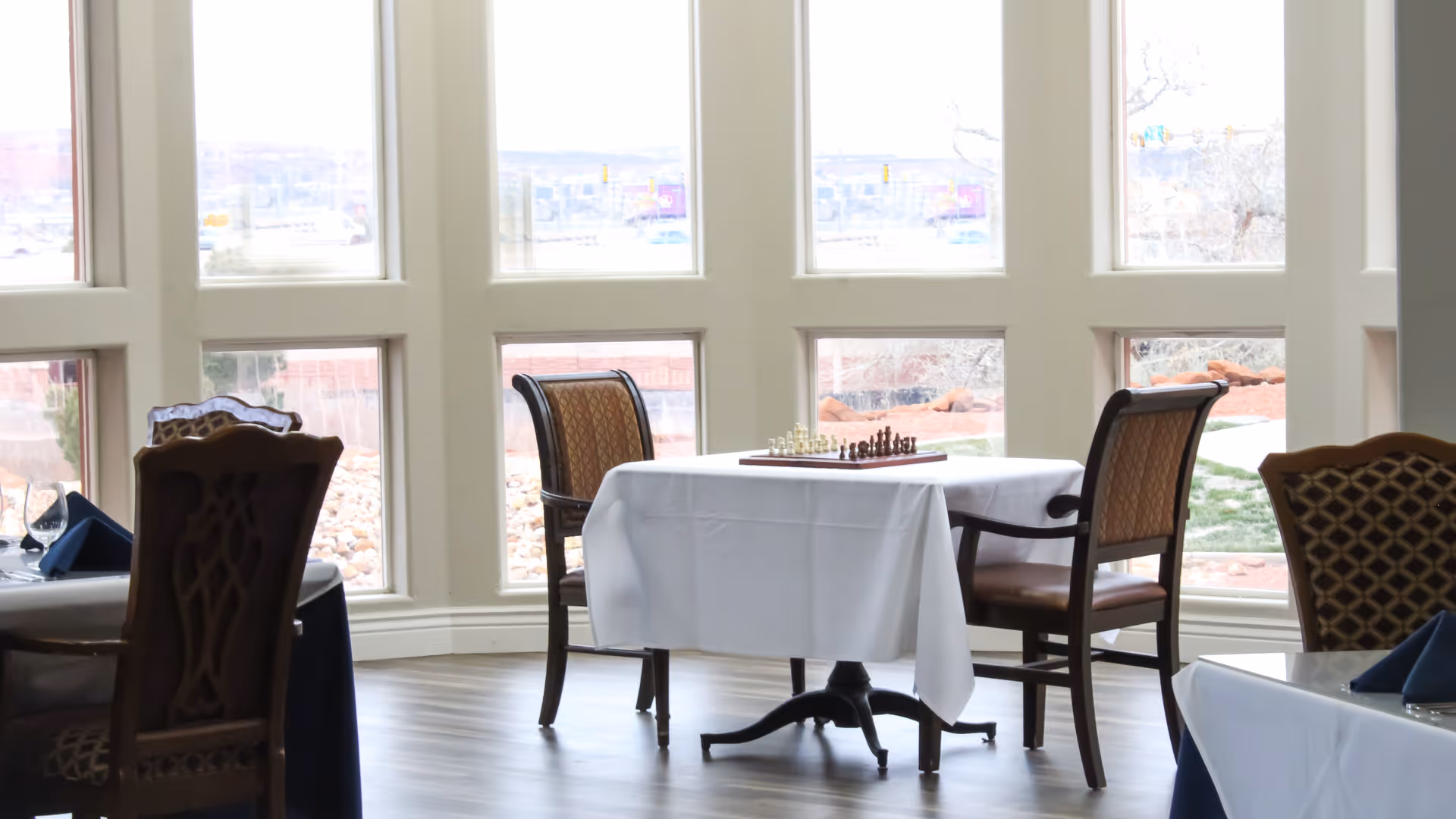 A dining area with large windows showing an outdoor view. There is a table covered with a white tablecloth and two chairs around it. On the table, there is a chessboard with chess pieces set up. Other tables with chairs and place settings are partially visible.
