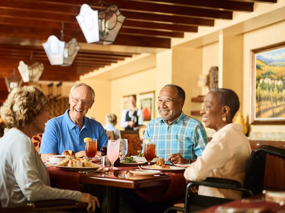 Four elderly people sitting around a dining table in a restaurant or dining room, smiling and enjoying a meal together. The table is set with plates of food and drinks, and the background shows warm lighting and framed artwork on the walls.
