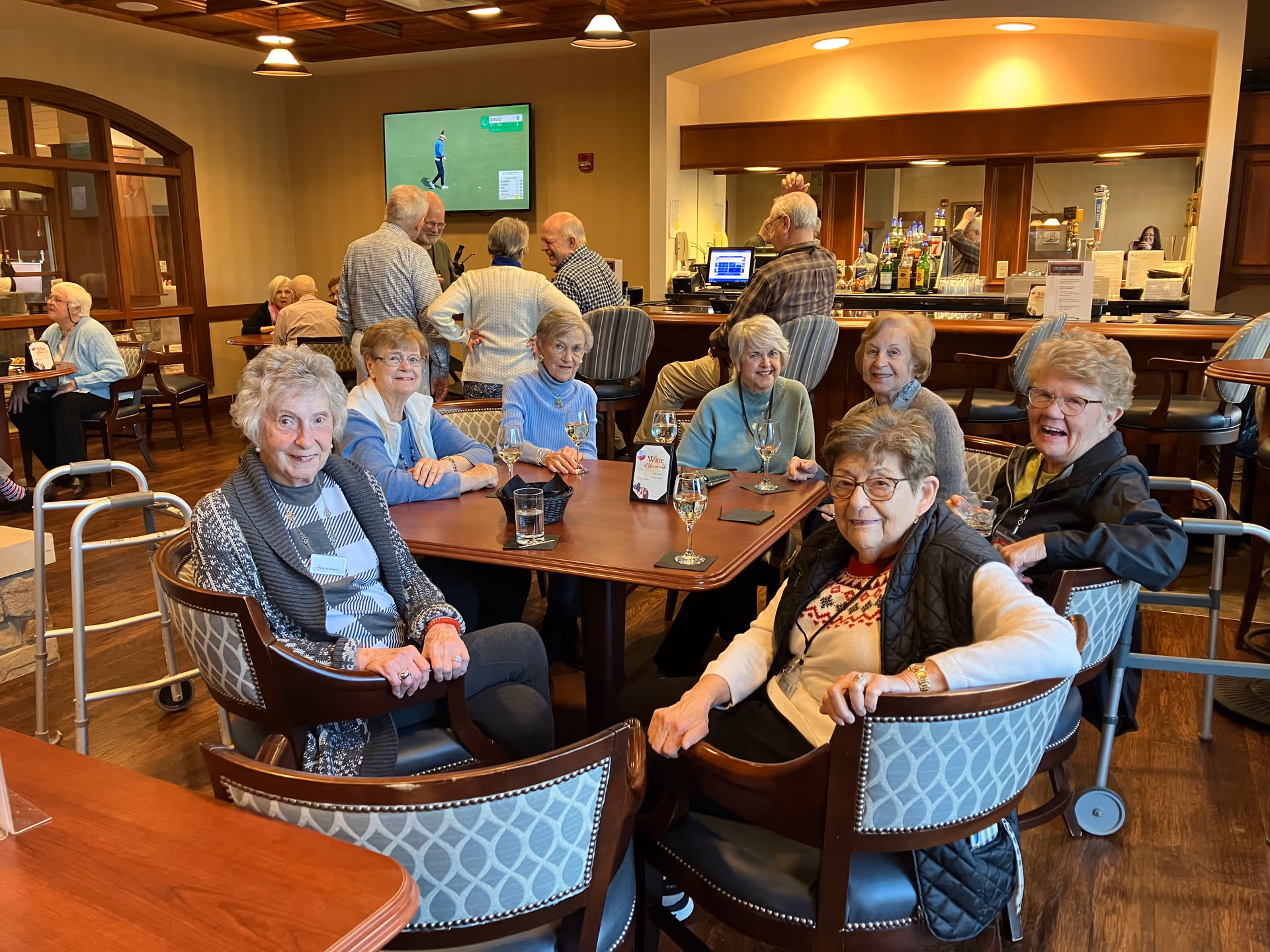 A group of elderly women sitting around a wooden table in a lounge area, smiling and enjoying drinks. In the background, other elderly people are standing and sitting near a bar with bottles and a TV mounted on the wall showing a golf game. The room has warm lighting and wooden furniture.