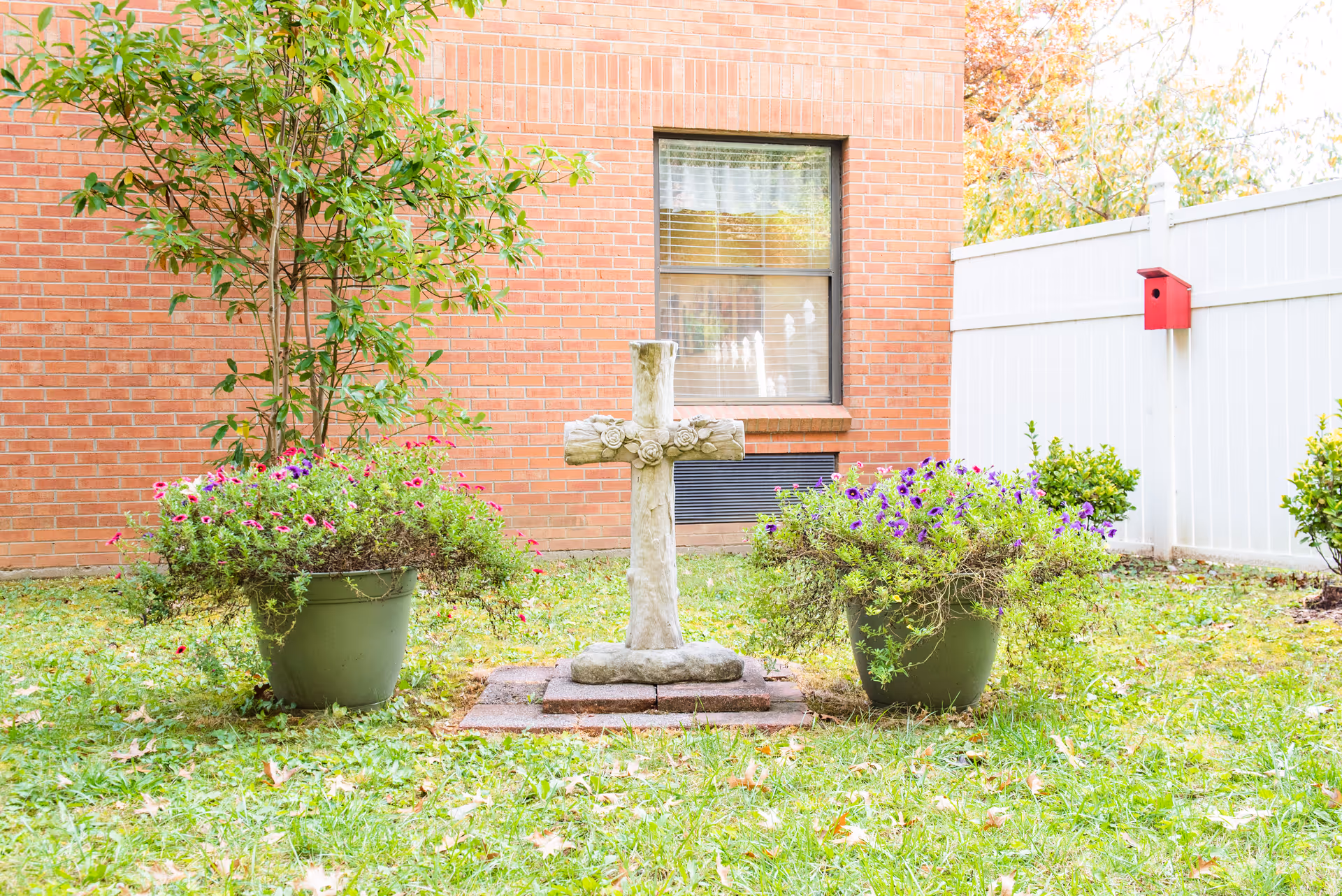 Stone cross monument flanked by two potted flowering plants on grass in a small courtyard with a brick building and white fence in the background.
