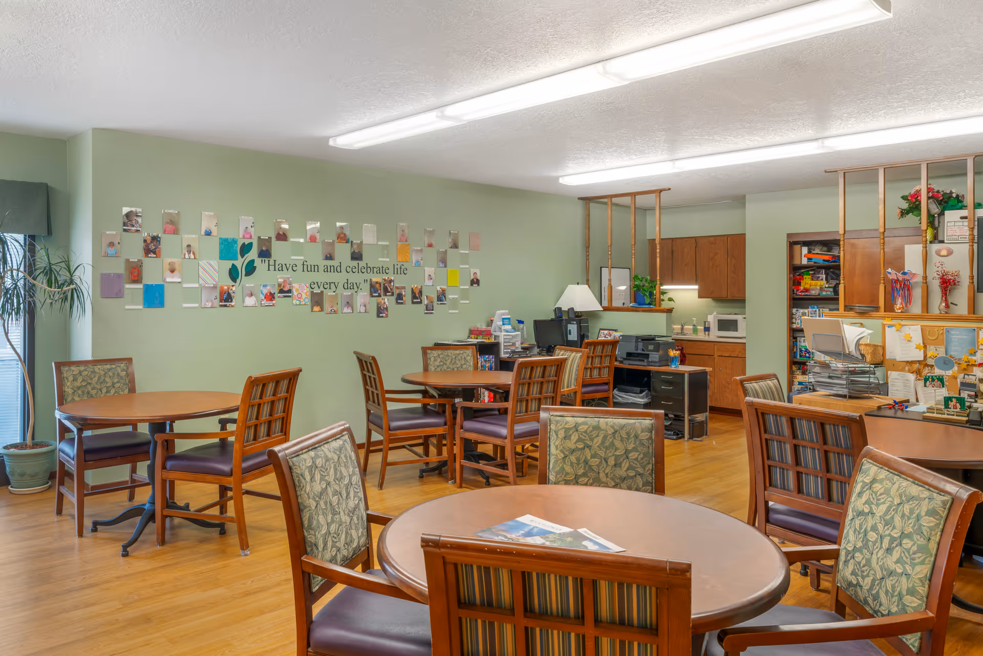 A cozy common area in a senior living facility with several round wooden tables and cushioned chairs arranged around the room. The walls are painted light green and decorated with photos and a quote that reads 'Have fun and celebrate life every day.' There is a small kitchenette area with wooden cabinets, a microwave, and a printer. The floor is wooden, and there is a potted plant near a window with a green valance.
