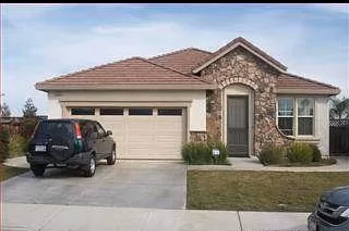 Front exterior view of a single-story house with a stone facade around the entrance, a two-car garage, a driveway with a parked black SUV, and a small lawn with shrubs.
