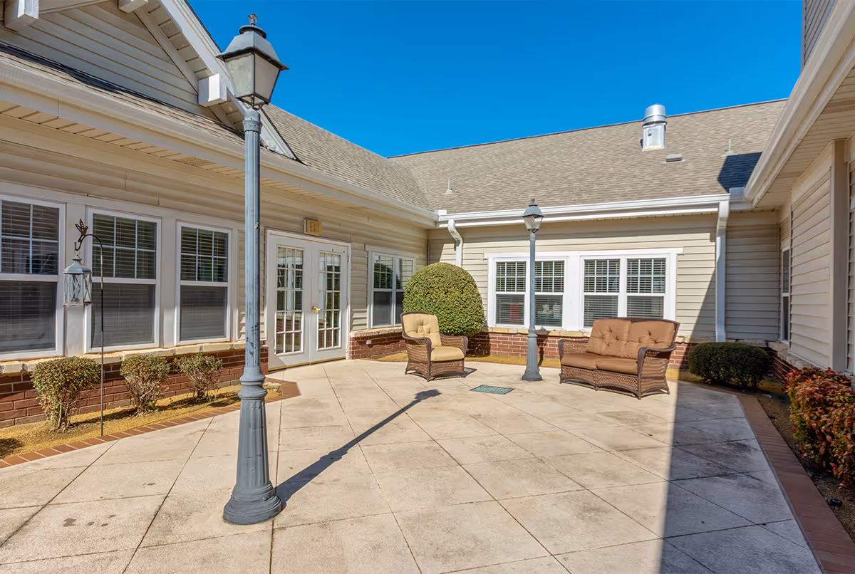 Outdoor courtyard area at Dorset Place Memory Care with two cushioned wicker chairs, two lamp posts, trimmed bushes, and beige siding building walls under a clear blue sky.