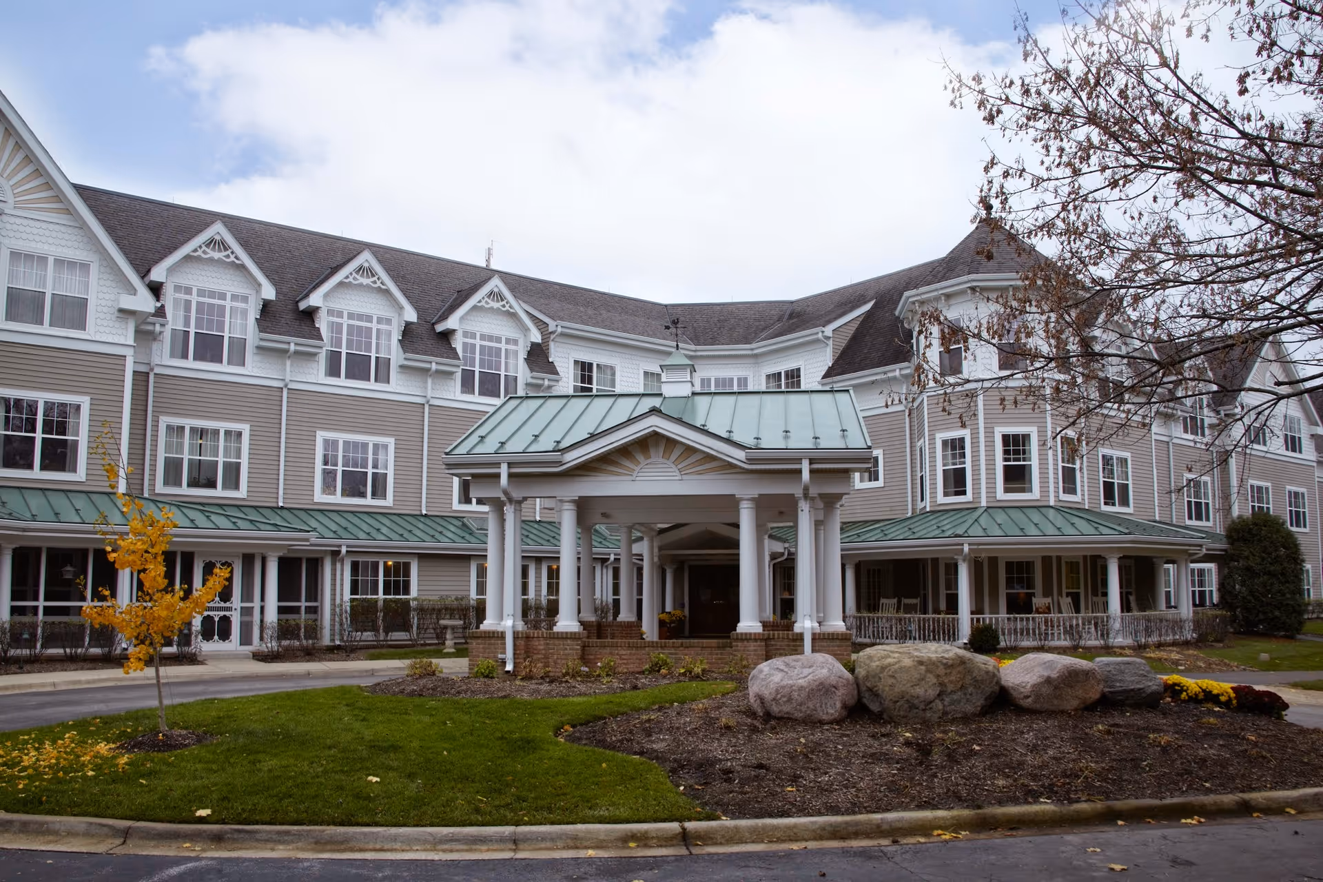 Exterior view of a senior living facility building with a covered entrance supported by white columns. The building has multiple windows, a green metal roof, and beige siding. There are some trees, bushes, and large rocks in the landscaped area in front of the entrance.
