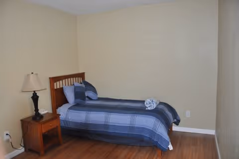 Simple bedroom with a twin bed against a beige wall, a wooden nightstand with a lamp, and hardwood floors.