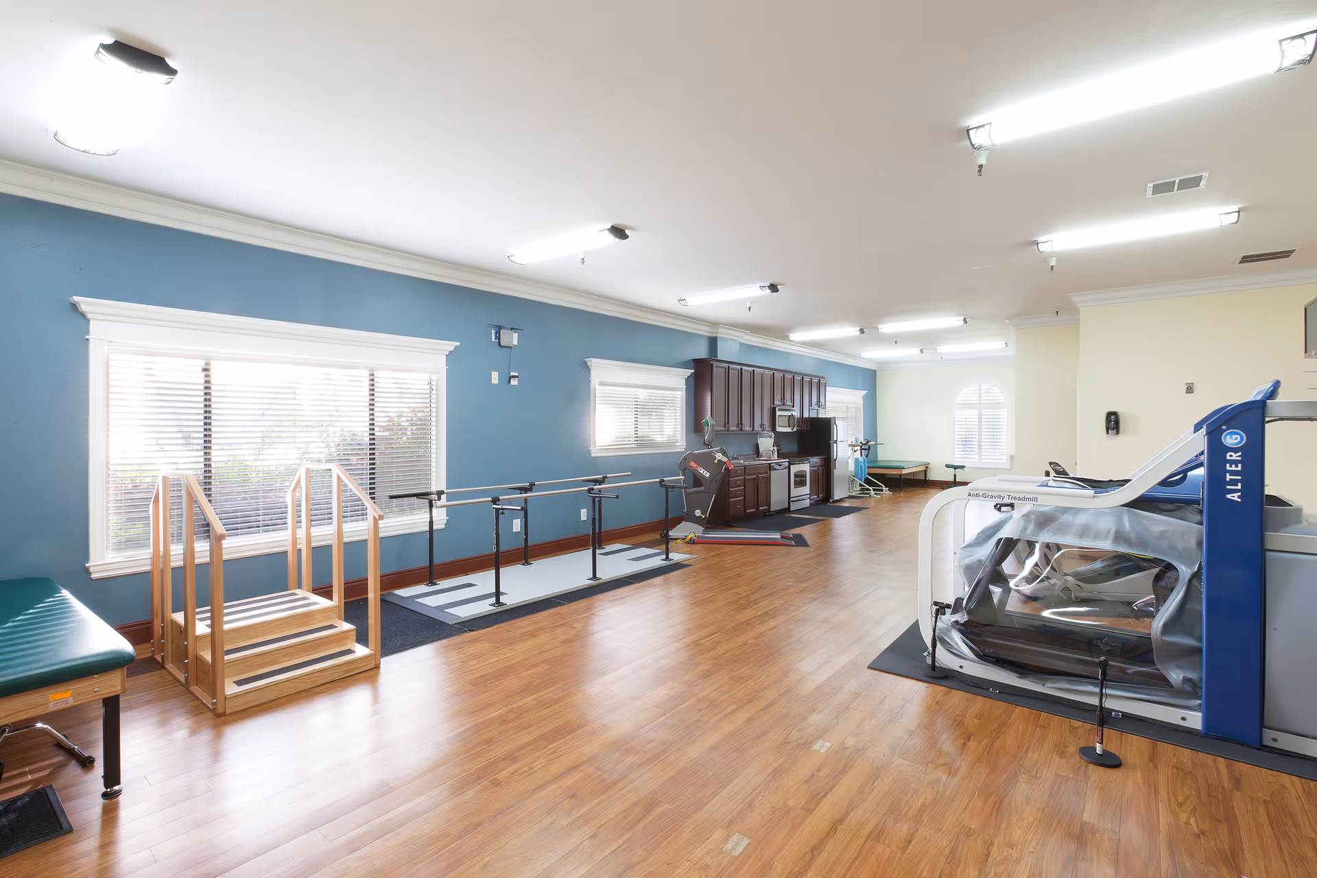 A spacious rehabilitation room with wooden flooring, blue and cream walls, and large windows with blinds. The room contains physical therapy equipment including parallel bars, a small wooden staircase, an anti-gravity treadmill, and a kitchenette area with cabinets, a microwave, and a refrigerator.
