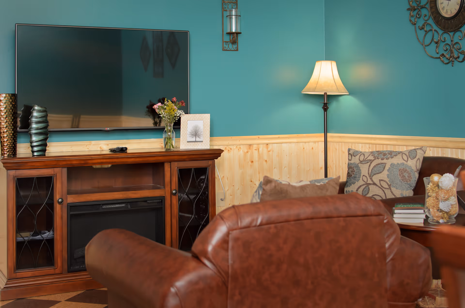 Cozy living room with teal walls and wood paneling, featuring a large flat-screen TV mounted above a wooden cabinet with decorative vases and a framed picture. In the foreground, there is a brown leather armchair and a matching sofa with patterned cushions. A floor lamp provides warm lighting, and a small side table holds books and a decorative glass container with pine cones.