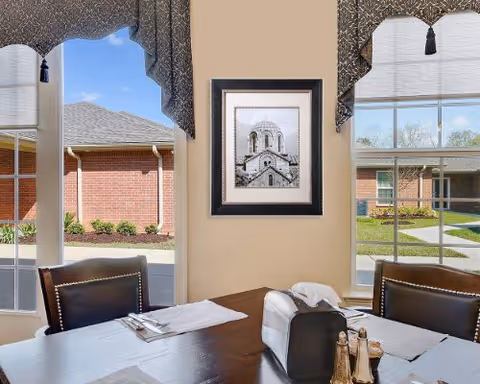 View from inside a dining area showing a wooden table set with placemats, napkins, and condiments. Two leather chairs are positioned at the table. Large windows with decorative valances provide a clear view of a courtyard with brick buildings and greenery outside. A framed black and white photo of a building is hung on the beige wall between the windows.