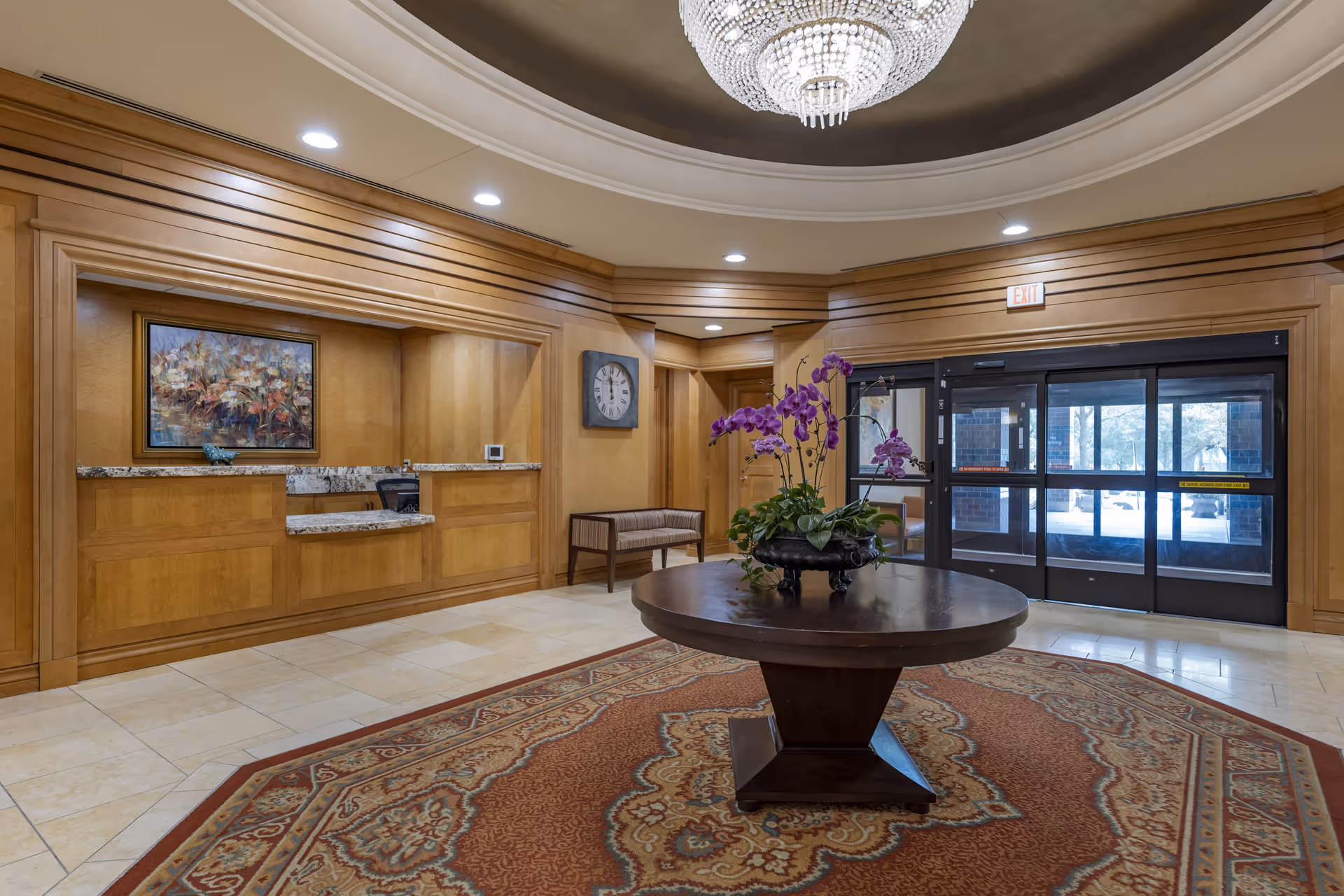 Lobby area with a round wooden table in the center holding a flower arrangement with purple orchids. The floor has a decorative rug, and the walls are paneled with wood. There is a reception desk with a granite countertop on the left side, a wall clock, a bench, and glass automatic doors leading outside. A large chandelier hangs from the ceiling.