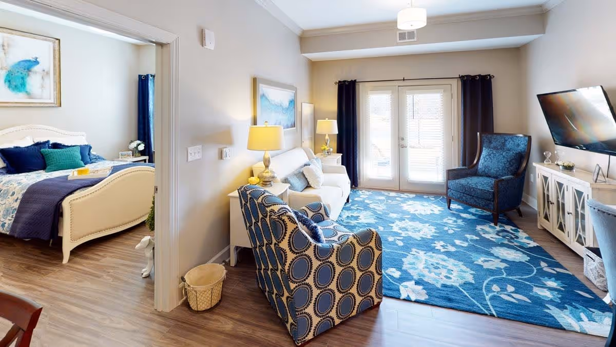 Bright furnished living room with patterned armchairs, a sofa, TV, and blue floral rug, with a bedroom visible through an open doorway.