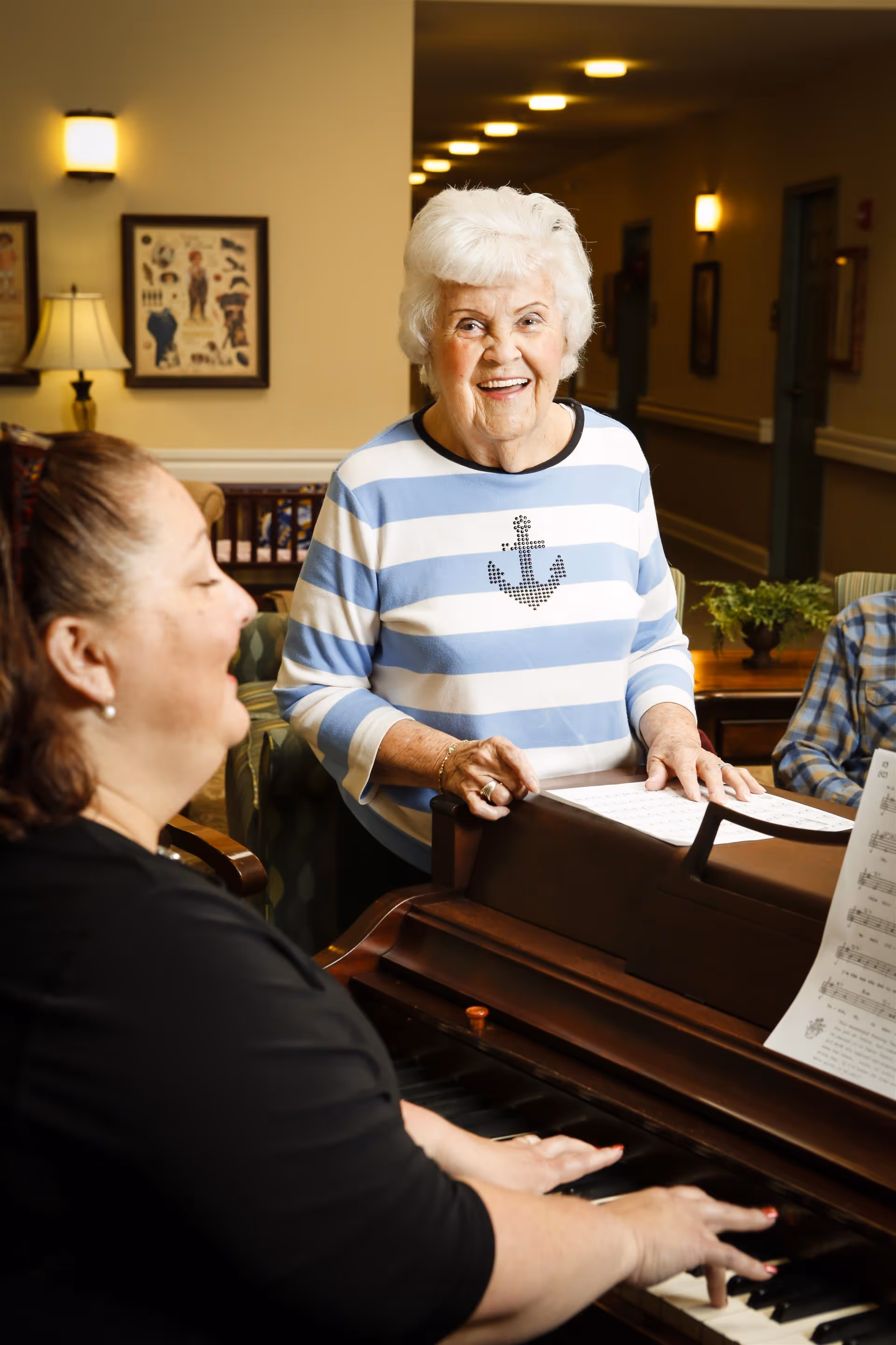 An elderly woman with white hair wearing a blue and white striped shirt with an anchor design stands smiling next to a piano. A woman with dark hair is playing the piano, and sheet music is visible on the piano. The setting appears to be an indoor common area with warm lighting and framed pictures on the wall.
