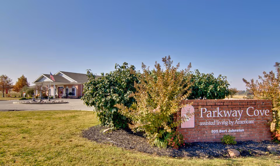Exterior view of Parkway Cove assisted living facility with a brick sign displaying the facility name and address, surrounded by bushes and trees, and a single-story building with an American flag in the background under a clear blue sky.