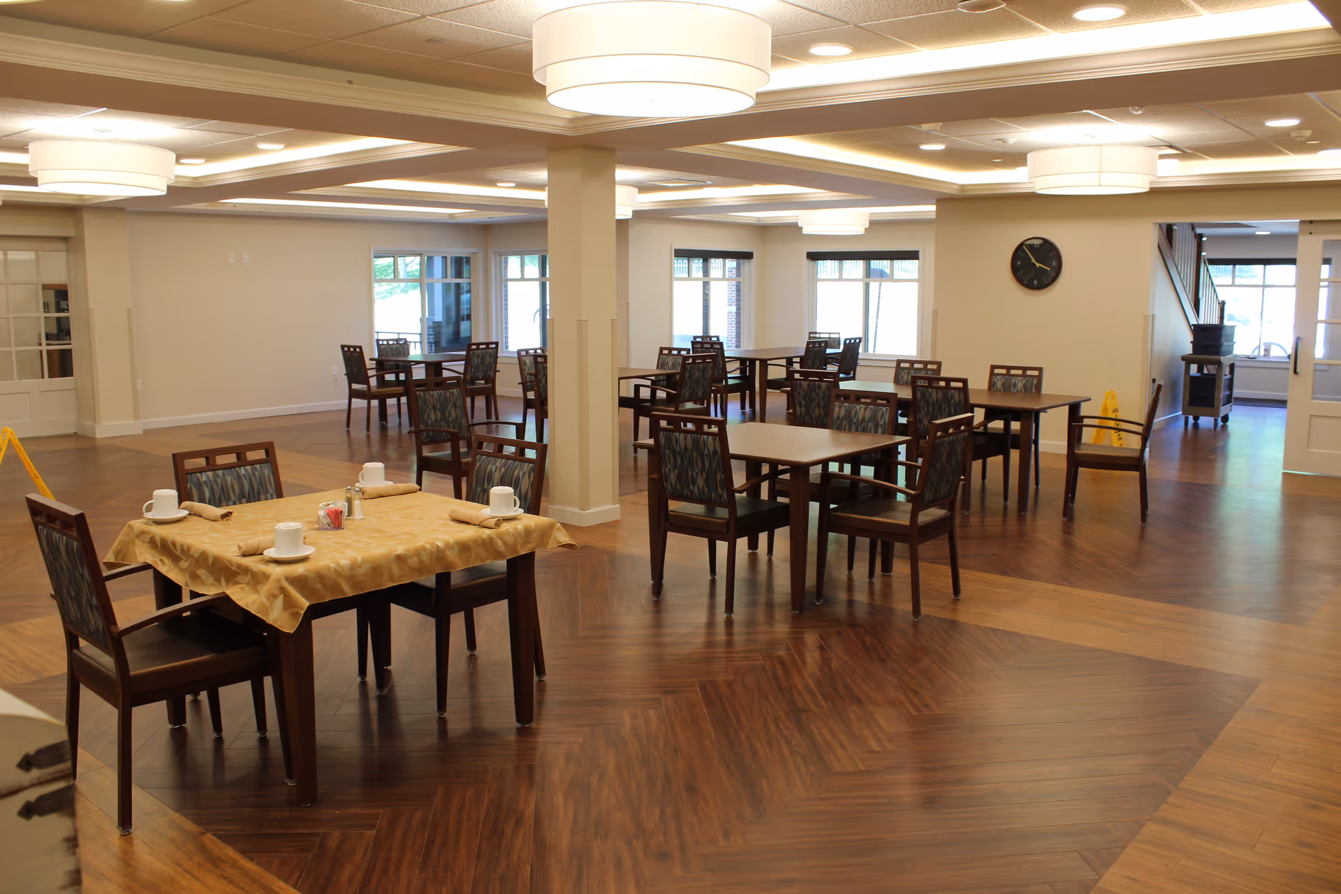 A spacious dining room with multiple wooden tables and chairs arranged neatly. One table in the foreground is covered with a beige tablecloth and set with cups, napkins, and condiments. The room has large windows allowing natural light, modern ceiling lights, and a wall clock. The floor is wooden with a patterned design.
