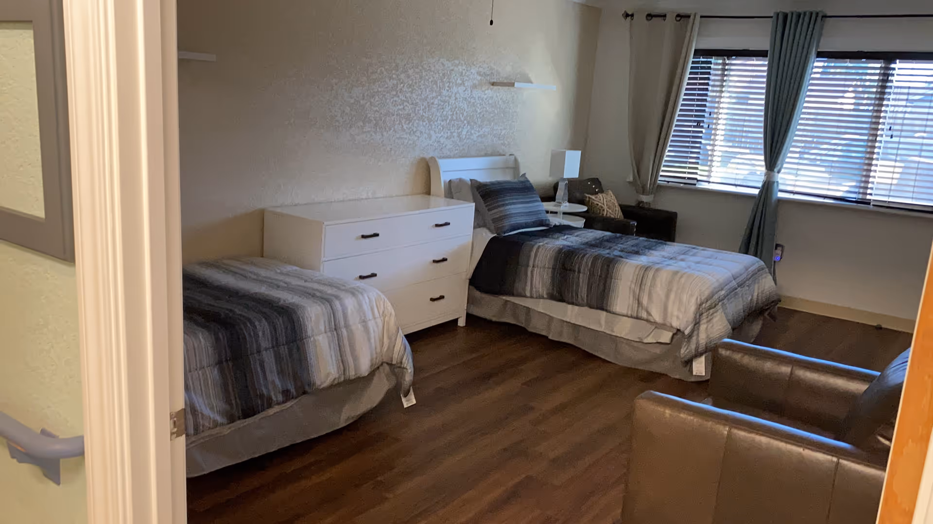 Shared bedroom with two twin beds, a white dresser, armchairs, and a window with blinds and curtains.